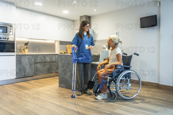 Professional caregiver providing home assistance to a senior woman in a wheelchair, discussing her health and well being in a modern kitchen with medical crutches nearby