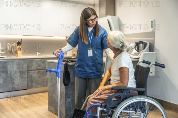 Young female caregiver in scrubs assisting a senior woman in a wheelchair, providing home healthcare support and discussing mobility with crutches in a modern kitchen setting