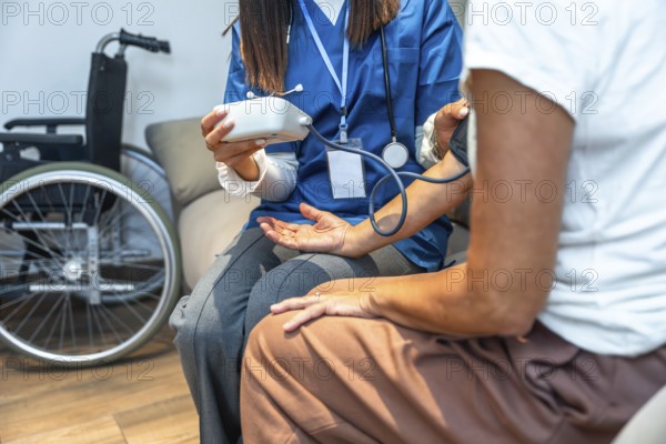 Healthcare professional checking a person's blood pressure with a cuff and digital monitor, providing medical consultation during a home visit with a wheelchair visible