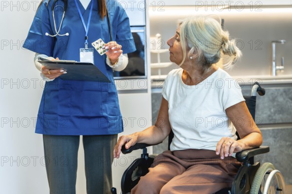 Healthcare professional wearing blue scrubs holding a clipboard and blister pack, explaining medication to a senior woman sitting in a wheelchair for home care assistance