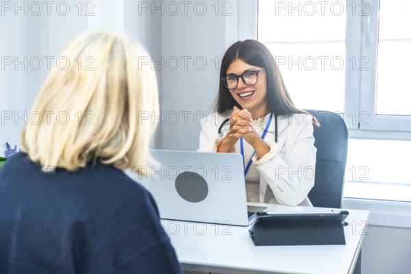 Young female doctor wearing a lab coat and stethoscope, smiling while actively listening to a patient during a healthcare consultation in a bright clinic office