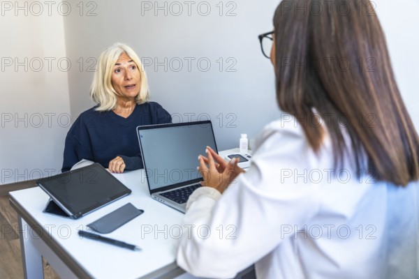 Senior woman is talking with a female doctor during a medical consultation in a modern office, discussing her health and treatment options with a laptop and medical equipment on the desk