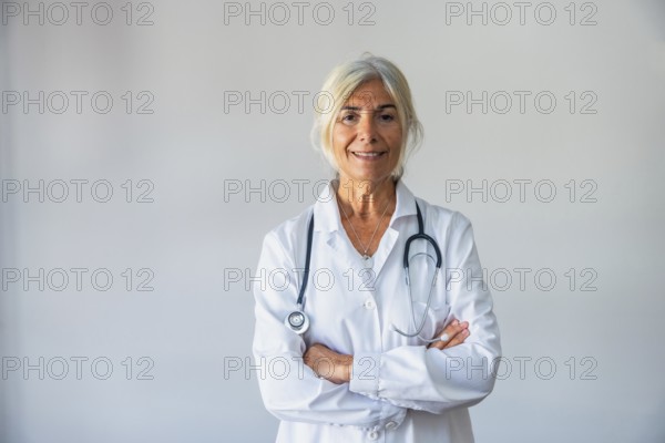 Senior female doctor with gray hair and a lab coat smiling confidently, standing against a neutral background with a stethoscope around her neck, representing healthcare and medical professionalism