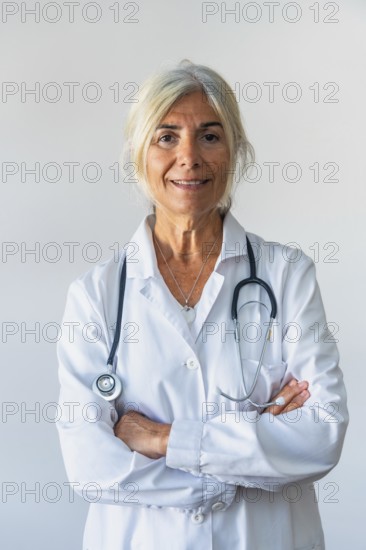 Senior woman doctor with gray hair and lab coat standing confidently with arms crossed, stethoscope around her neck and smiling at camera, conveying experience, trust and professional medical care
