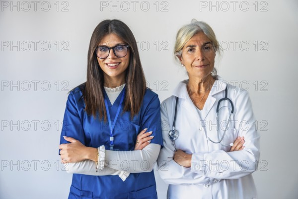 Two women healthcare professionals, a younger nurse in blue scrubs and an older doctor in a white coat with a stethoscope, standing confidently with folded arms against a white background