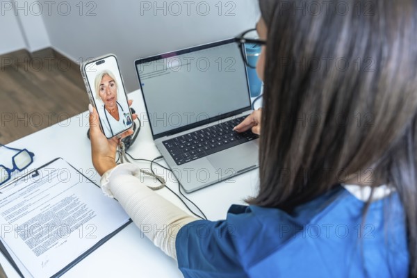 Healthcare professional having a video conference with a doctor on a smartphone while using a laptop, representing modern telehealth services and digital medical communication