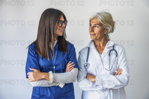 Two confident female doctors, one younger and one older, standing with crossed arms against a white background, representing teamwork and healthcare professionalism