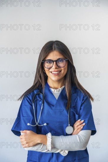Young woman doctor wearing blue scrubs, a stethoscope, and eyeglasses, standing confidently with arms crossed, smiling at the camera representing healthcare and medicine