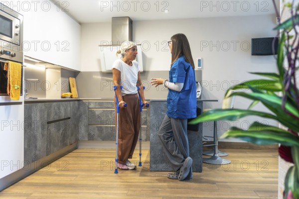 Home caregiver interacting with a senior woman using crutches, providing support and advice for her recovery and rehabilitation process in a modern kitchen setting