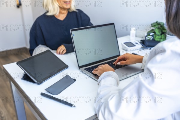 Doctor engaging in a medical consultation with a senior patient, typing health information and records on a laptop computer during an appointment at a clinic desk