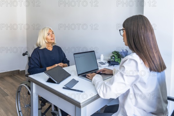 Female doctor consulting with an older woman in a wheelchair, discussing treatment and care while reviewing health information on a laptop and tablet in a clinic office setting