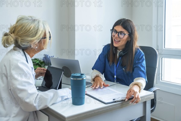 Healthcare professionals, a doctor in a lab coat and a nurse in scrubs, sitting at a desk and discussing medical information during a consultation using digital devices in a clinic office