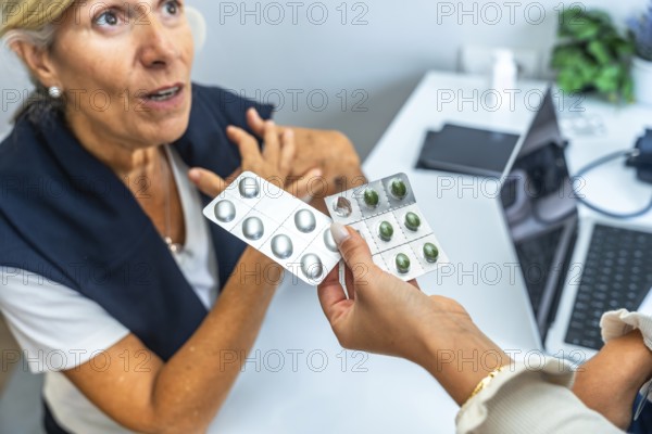Doctor hand holding two blister packs of pills, one with green capsules and one with silver capsules, offering medicine to a concerned senior woman discussing health issues during a consultation