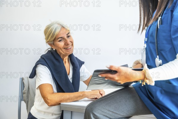 Senior woman smiling and listening as doctor in blue scrubs reviews a medical chart, sharing a compassionate consultation about health, care, and wellness in a clinic setting