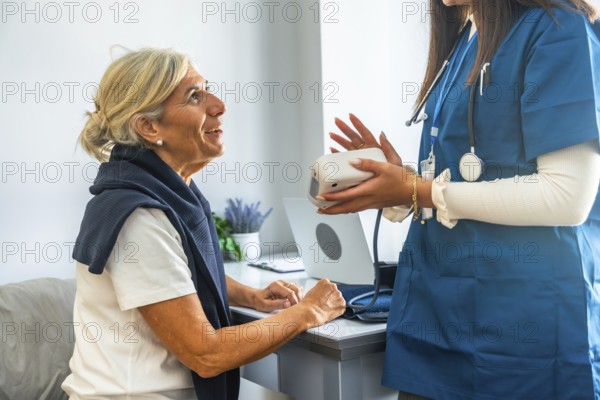 Healthcare professional in blue scrubs holding a blood pressure monitor, explaining its use to an attentive senior woman during a medical consultation