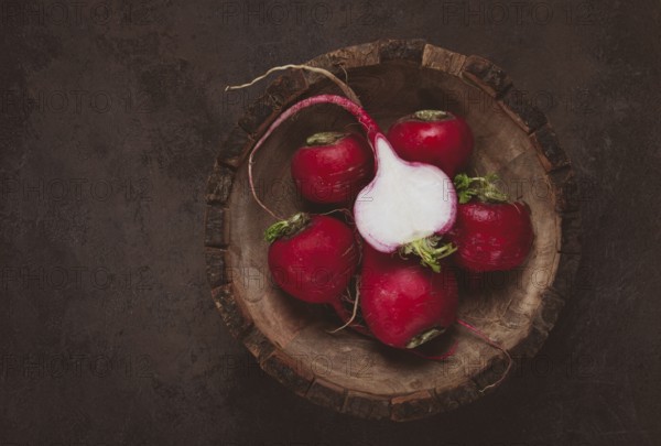 A collection of vibrant red radishes sits in a carved wooden bowl on a dark, textured surface. The fresh vegetables are perfect for salads or garnishes, showcasing their natural beauty