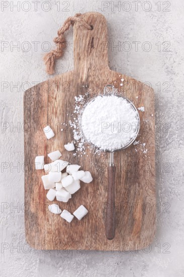 A strainer with powdered sugar, on a wooden cutting board, with pieces of white sugar