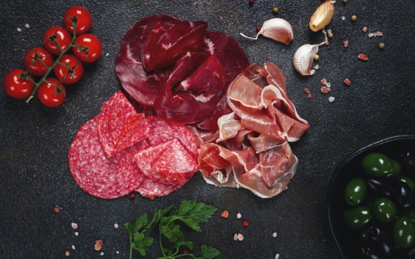 Assorted dried meat, fresh tomatoes and olives spread out on a dark surface, food composition, top view, no people