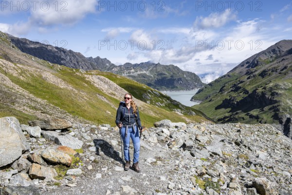 Person posing on rocky path with background of mountains and a lake under blue sky, Lac des Dix reservoir, Valais Alps, Valais, Switzerland