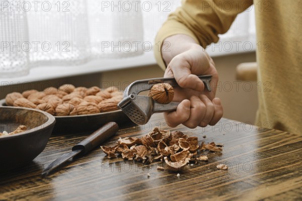 A person is breaking open a walnut with a metal nutcracker, sitting at a wooden table. Shelled nuts are scattered around, creating a warm and inviting atmosphere in the kitchen during daylight hours