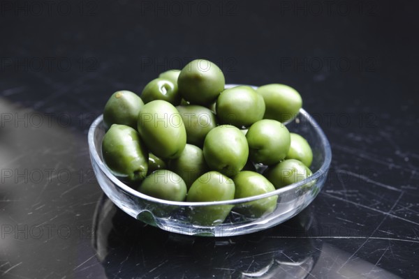 A clear bowl holds a generous pile of green olives. The olives are glossy and vibrant, resting on a dark surface that enhances their bright color