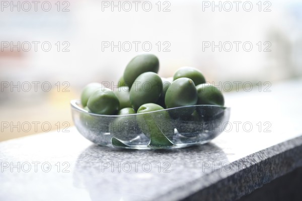 A bowl filled with green olives on a smooth granite surface. The sunlight reflects off the glass while the vibrant olives appear inviting against a blurred urban background