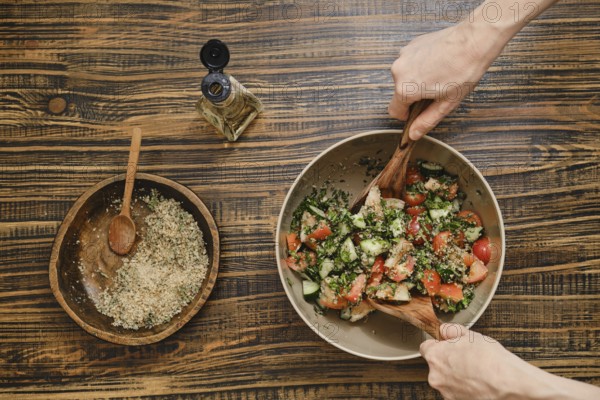 Two hands mix fresh vegetables in a bowl while preparing a salad. Nearby, a smaller bowl holds seasoning and garnishing ingredients, creating a vibrant and tasty dish