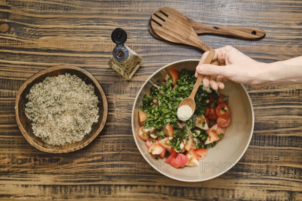 A hand adds seasoning to a bowl filled with chopped tomatoes and fresh herbs in a cozy kitchen setting