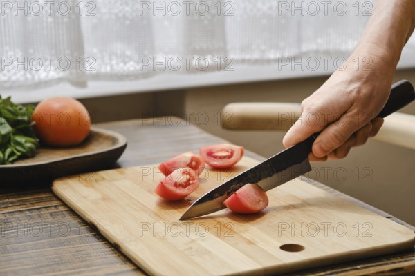 A person is slicing ripe tomatoes on a wooden cutting board with a sharp knife. Bright sunlight streams through the window, illuminating fresh ingredients on a kitchen countertop
