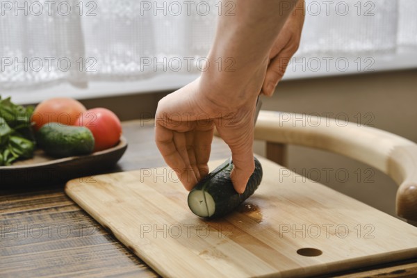 A hand holds a cucumber while cutting it on a wooden board. Fresh tomatoes and greens are arranged on a plate beside it, suggesting a meal preparation. Sunlight shines through the window