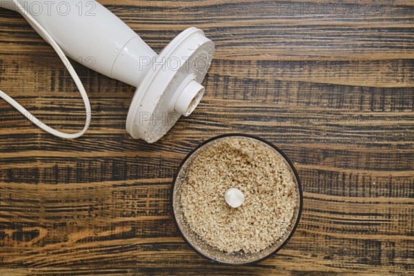 A food processor on a wooden countertop, with finely ground walnuts in the mixing bowl. The scene shows kitchen preparation, ready for cooking or baking tasks
