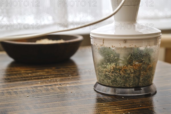 Chopping fresh herbs and nuts in a food processor near a wooden bowl of grains on a kitchen table