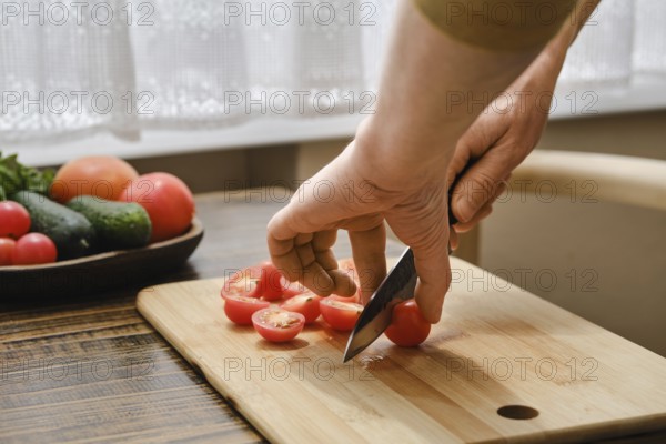 A person is skillfully slicing ripe cherry tomatoes on a wooden cutting board in a bright kitchen. Fresh vegetables are laid out nearby, showcasing the ingredients for a healthy meal