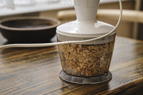 Chopping walnuts in a food processor on a rustic wooden table. The device is plugged in and the chopped mixture is visible in its container, surrounded by a cozy kitchen setting