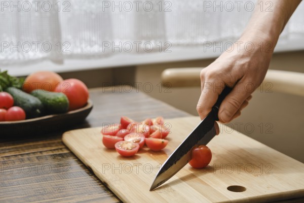 A hand holds a knife, skillfully slicing fresh cherry tomatoes on a wooden cutting board. Various vegetables are visible in the background