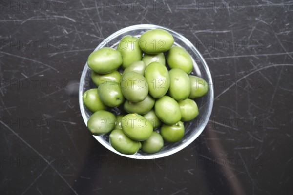 Overhead view of a bowl filled with green olives on a black surface. The olives are vibrant and glossy, showcasing their natural color