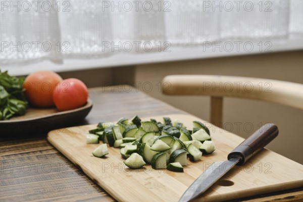 Freshly chopped cucumbers on a wooden cutting board beside a knife. A bowl of tomatoes is visible in the background. Natural light filters through a window, giving a cozy kitchen vibe