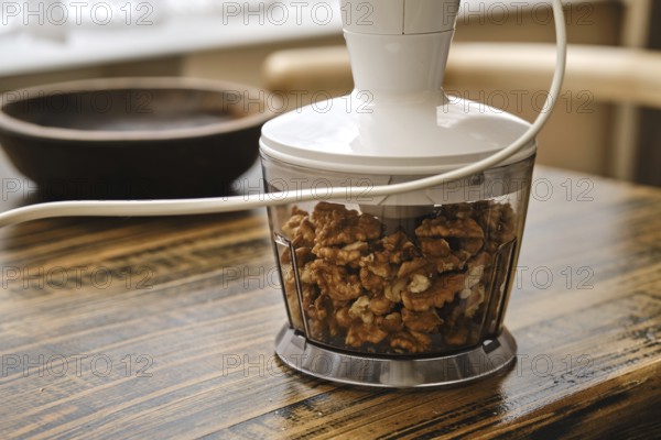 A food processor is filled with walnuts on a wooden table. Natural light filters through a nearby window, creating a cozy kitchen atmosphere