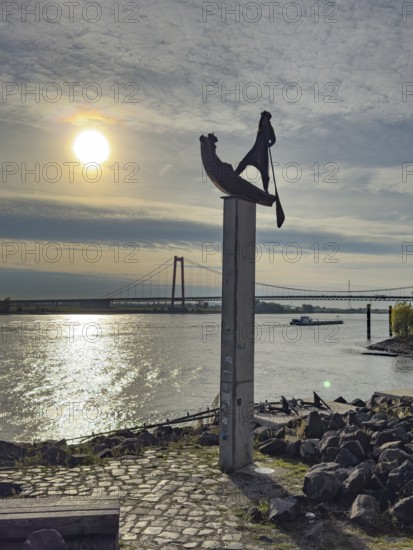 Photo with melancholy atmosphere of sculpture Fährmann Artwork on pillar stele by sculptor Dieter von Levetzow from 1983 on the Rhine promenade in Emmerich, reminiscence of time in front of the Rhine bridge, on the river right under Rhine bridge Freight ship freighter for inland navigation, Emmerich, Lower Rhine, North Rhine-Westphalia, Germany