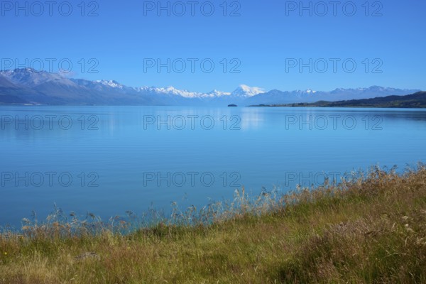View over grassy banks to the mountain range behind the lake, Lake Pukaki Viewpoint, Pukaki, Canterbury, South Island, New Zealand