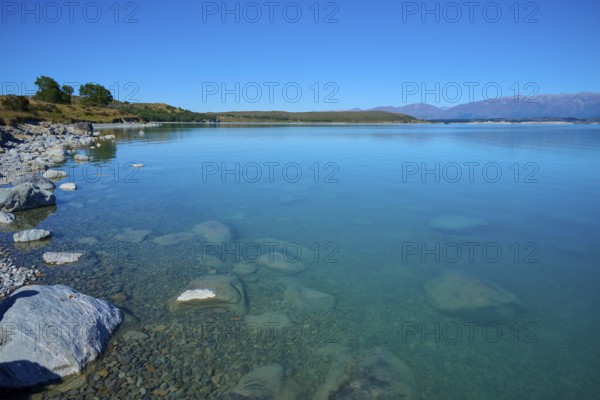 Shore with stones in clear water surrounded by vast countryside and mountains, Lake Pukaki Viewpoint, Pukaki, Canterbury, South Island, New Zealand