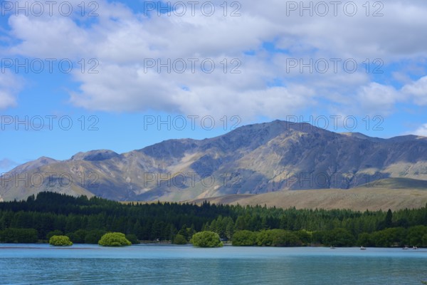 Mountainous landscape with a lake in the foreground and wooded hills under a cloudy sky, Lake Tekapo, Canterbury, South Island, New Zealand