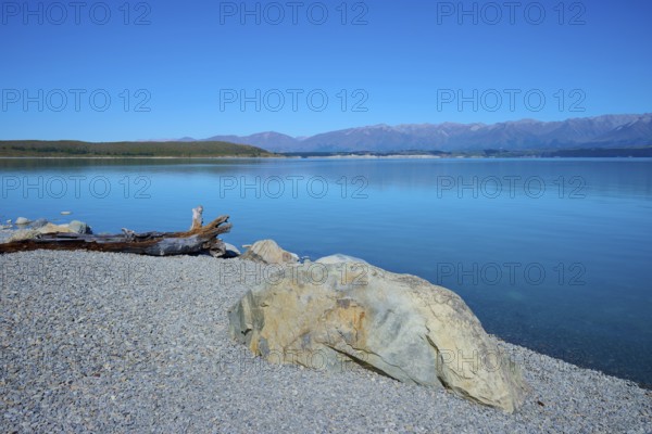 Rocky shore on a calm blue lake with views of distant mountains, Lake Pukaki Viewpoint, Pukaki, Canterbury, South Island, New Zealand