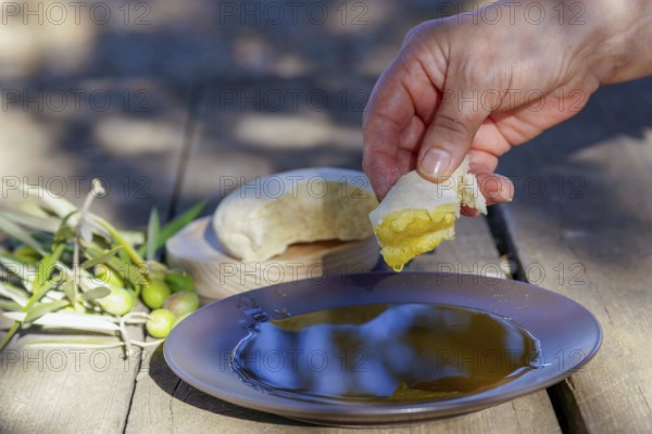 Hand dipping fresh bread into a plate of extra virgin olive oil on a rustic wooden table, with olives and olive branches