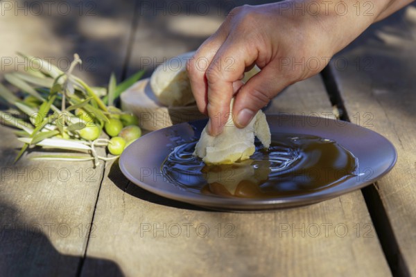 Hand dipping a piece of fresh bread into a shallow plate of extra virgin olive oil, with olives and olive branches on a rustic wooden table