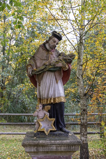 Religious statue with cape and angel in an autumnal park in front of yellow leaves, Weinviertel Lower Austria