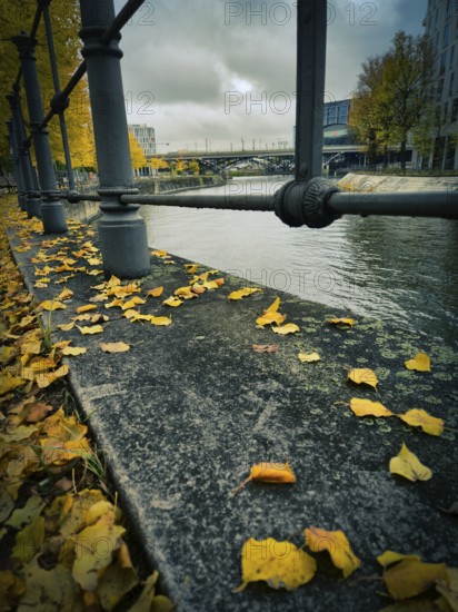 Wet stone edge on the canal with yellow autumn leaves and solid railings, in the background bridge and city under heavy cloudy sky, at Berlin Central Station, 10557 Berlin, Germany