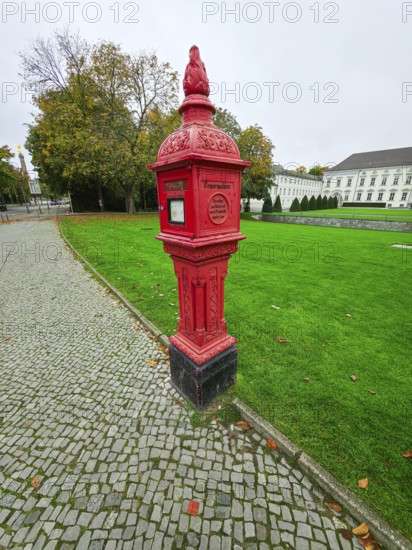 Historic red fire alarm column on cobblestones on the roadside of the Federal President's office, green lawn and cloudy sky, quiet autumn atmosphere, at Bellevue Palace in 10557 Berlin, Germany
