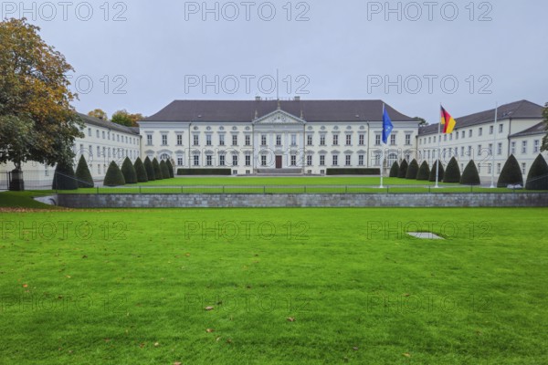 Neoclassical castle in front of a well-kept lawn, lined with topiaries, under grey skies with waving flags and a quiet autumn atmosphere, at Bellevue Palace in 10557 Berlin, Germany