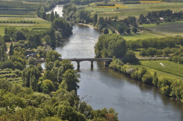 An aerial view of a serene river flowing under a bridge, surrounded by lush green fields and trees. Dordogne seen from Domme in Périgord, France